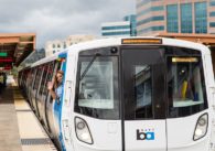 Woman waving out the driver's window of a new BART train while people wait on the platform
