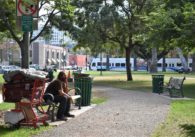 A homeless man sits on a bench in a park, with his cart of stuff nearby