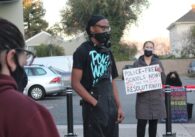 A person in overstays stands outside wearing a face mask, with another person behind them holding a sign