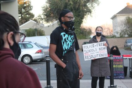 A person in overstays stands outside wearing a face mask, with another person behind them holding a sign
