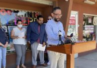 A man stands at a podium outside with people in the background