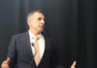 A man in a suit and tie stands at a microphone in front of a black backdrop