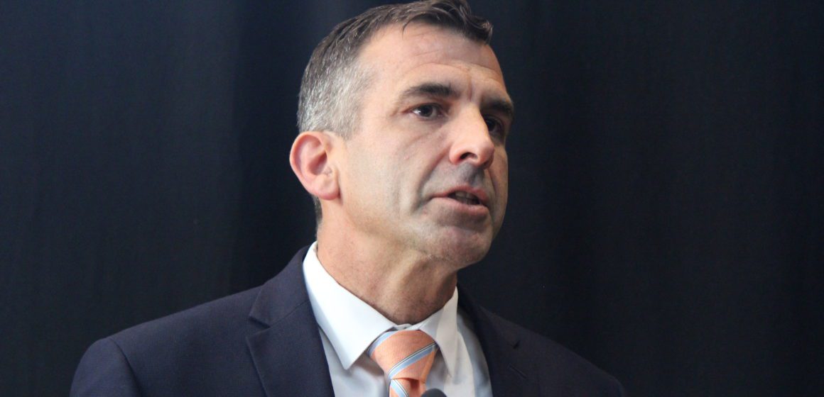 A man in a suit speaks into a microphone in front of a black backdrop