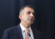 A man in a suit speaks into a microphone in front of a black backdrop