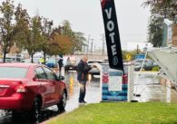 A man standing in the rain next to a car puts a mail-in ballot into a drop box