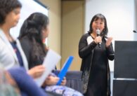 A woman speaks into a microphone, addressing other people seated nearby