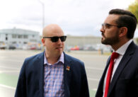 A man in sunglasses and a blazer stands next to a man in a suit