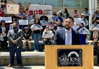 A man standing outside at a podium with a crowd of people behind him
