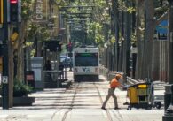 A man pushes a cart across the street as a light rail train moves in the background