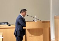 A man in a suit speaks into a microphone at a podium