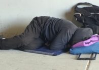 A homeless person sleeps on the ground under a highway overpass