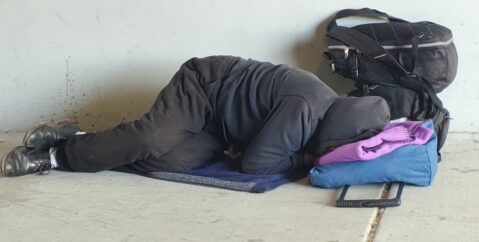 A homeless person sleeps on the ground under a highway overpass