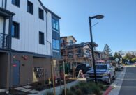 A finished white multi-story house next to a wooden house still under construction with scaffolding. In front of the houses is greenery and a pickup truck.