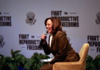 A woman sitting in a chair speaks into a microphone onstage.