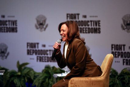 A woman sitting in a chair speaks into a microphone onstage.