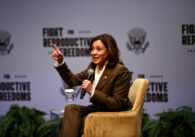 A woman sitting in a chair speaks into a microphone onstage.