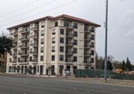 A six-story beige apartment building with a terracotta-colored roof sits behind an asphalt road.