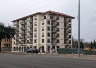 A six-story beige apartment building with a terracotta-colored roof sits behind an asphalt road.