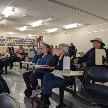 Un grupo de personas se sientan mirando hacia adelante en un salón de clases en escritorios escalonados. Luces fluorescentes se alinean en el techo.