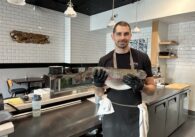A man holding a fish stands behind the counter in a restaurant.