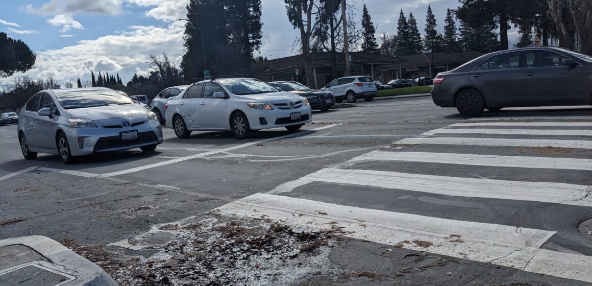 Cars drive through an intersection, behind a crosswalk under a cloudy blue sky.