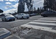 Cars drive through an intersection, behind a crosswalk under a cloudy blue sky.