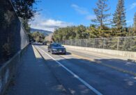 A black sedan drives across the Blossom Hill Road bridge, with cars going in both directions.