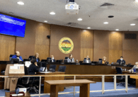 Seven people sitting on elevated dais with people working on desks in front of them