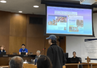 Person standing at podium wearing baseball cap with projection presentation of screenshotted Facebook ads