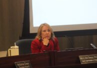 A woman sits at a dais in front of a projector screen at a school board meeting.