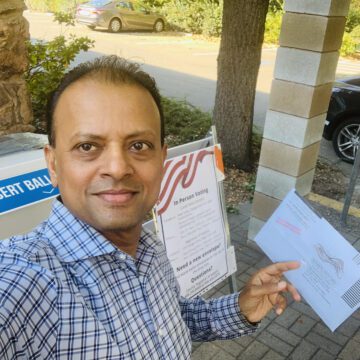 Rishi Kumar voting A man stands outside near an election ballot drop box holding a mail-in ballot.