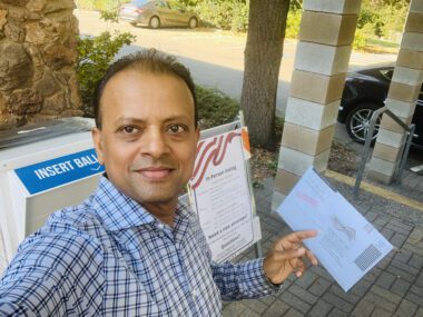 Rishi Kumar voting A man stands outside near an election ballot drop box holding a mail-in ballot.
