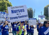 Group of striking nurses all wearing blue shirts reading #RNPASTRONG and holding signs, foreground nurse holding sign that reads" Santa Clara County Nurses for a Strong Contract"