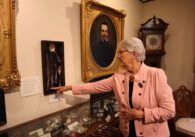 White haired woman in pink blazer pointing at silver cutlery mounted on museum wall, beside oil portrait and antique grandfather clock