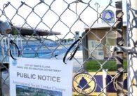 Chainlink fence with posted notice reading "City of Santa Clara Parks and Recreation Department Public Notice International Swim Center Closure." Behind the fence is the Santa Clara Swim Club notice board and two large pools with an audience section.