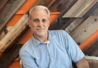 A man sitting in front of a wooden backdrop stares at the camera