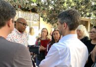 Chuck Cantrell, a Black man wearing thick rimmed glasses and a floral button down shirt, talking to Matt Mahan, a Caucasian man with short hair and a goatee, while surrounded by a group of people