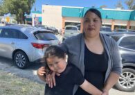 A woman stands outside in front of cars with her young son