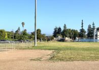Mostly empty baseball field and lawn with some people standing on side