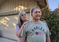 An older woman stands behind an older man, putting her hands on his shoulders
