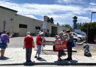 Group of people holding signs and standing on sidewalk near homeless shelter parking lot, with signs reading "60% of the 104 unhoused deaths — Black/Brown Seniors/Singles" and "Hot Senior, Let us in"