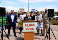 Người phụ nữ tóc vàng mặc áo sơ mi denim đứng sau bục phát biểu với biển hiệu 75,000 việc làm, trước nhóm người có biển hiệu "Sẵn sàng làm việc"