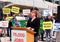 Brunette woman with black glasses in a black shirt speaking at podium reading "75,000 jobs" with group of union workers holding signs behind her reading "Ready to work," "Shovel Ready" and "75,000 jobs"