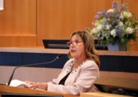Woman with curled brunette hair and red glasses speaking at a podium with flowers behind her