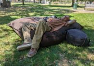 An unhoused man lies on a large brown cushion in the grass