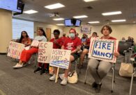 A row of people sitting in chairs holding protest signs at a school board meeting