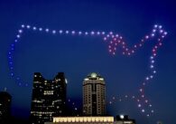 Dozens of drones glowing different colors glowing and forming the shape of the United States over buildings