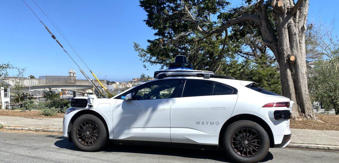 White car with mounted cameras labeled "Waymo" sitting by side of road