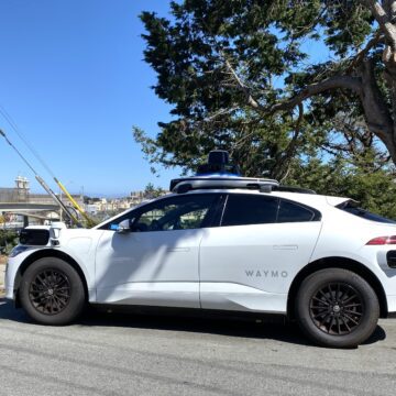 White car with mounted cameras labeled "Waymo" sitting by side of road