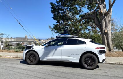 White car with mounted cameras labeled "Waymo" sitting by side of road
